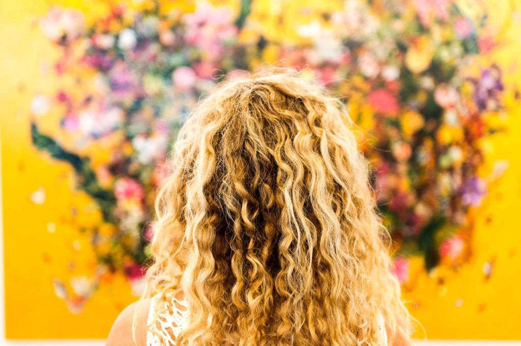 A back view of a woman with long, curly blonde hair, standing in a colorful, flower-filled environment.