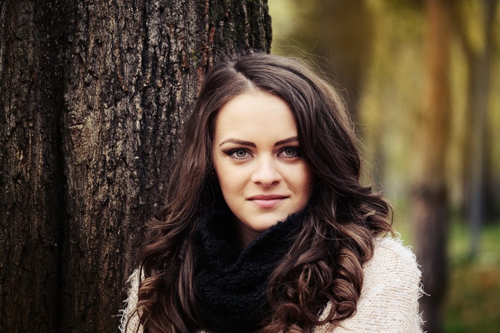 A woman with dark wavy hair, leaning against a tree outdoors.