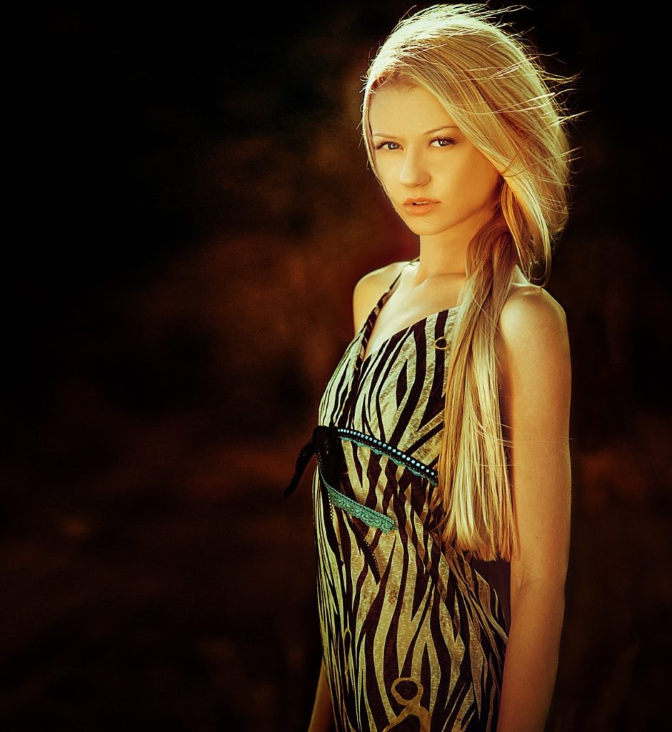 A blonde woman with long straight hair, posing in a dimly lit environment.