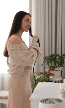 A woman in a cozy sweater, standing in front of a mirror, straightening her hair.